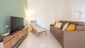 A living room with a flat-screen TV on a wooden console, a beige armchair, and a brown sofa with patterned cushions. - Sleek City Oasis - City Centre (Cagliari)