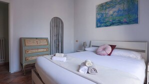 A neatly made bed with rolled towels, a dresser with decorative panels, a painting on the wall, and a radiator. - La Dolce Vita - IRIDE's Nest (Roma)