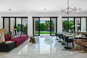 A modern interior with a dining area, a sofa, and large windows offering a view of a pool and greenery.
