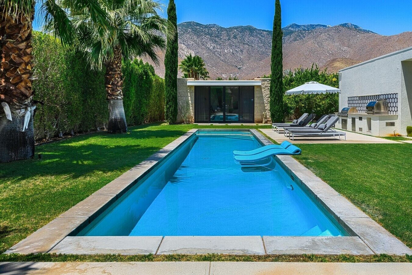 A rectangular swimming pool with a clear blue water, surrounded by a stone border. There are lounge chairs and a covered area with a table and chairs.