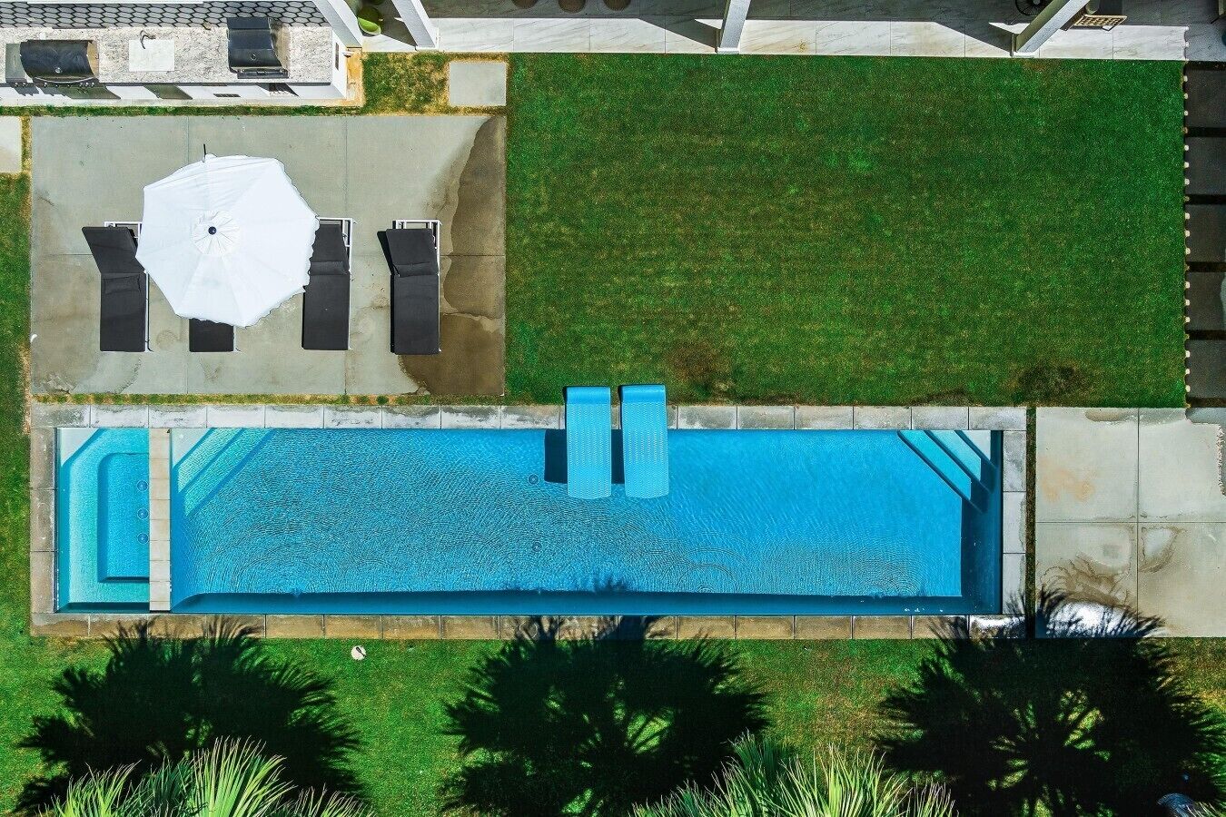 Ovenudblik over en boligejendom med swimmingpool, terrasse med bord og stole, samt frodig beplantning omkring.