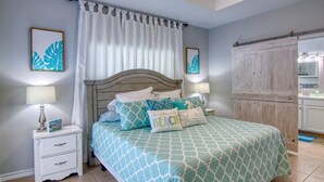 Une chambre à coucher avec un lit, deux tables de chevet, une commode, une armoire en bois et une salle de bain visible à travers une porte ouverte.