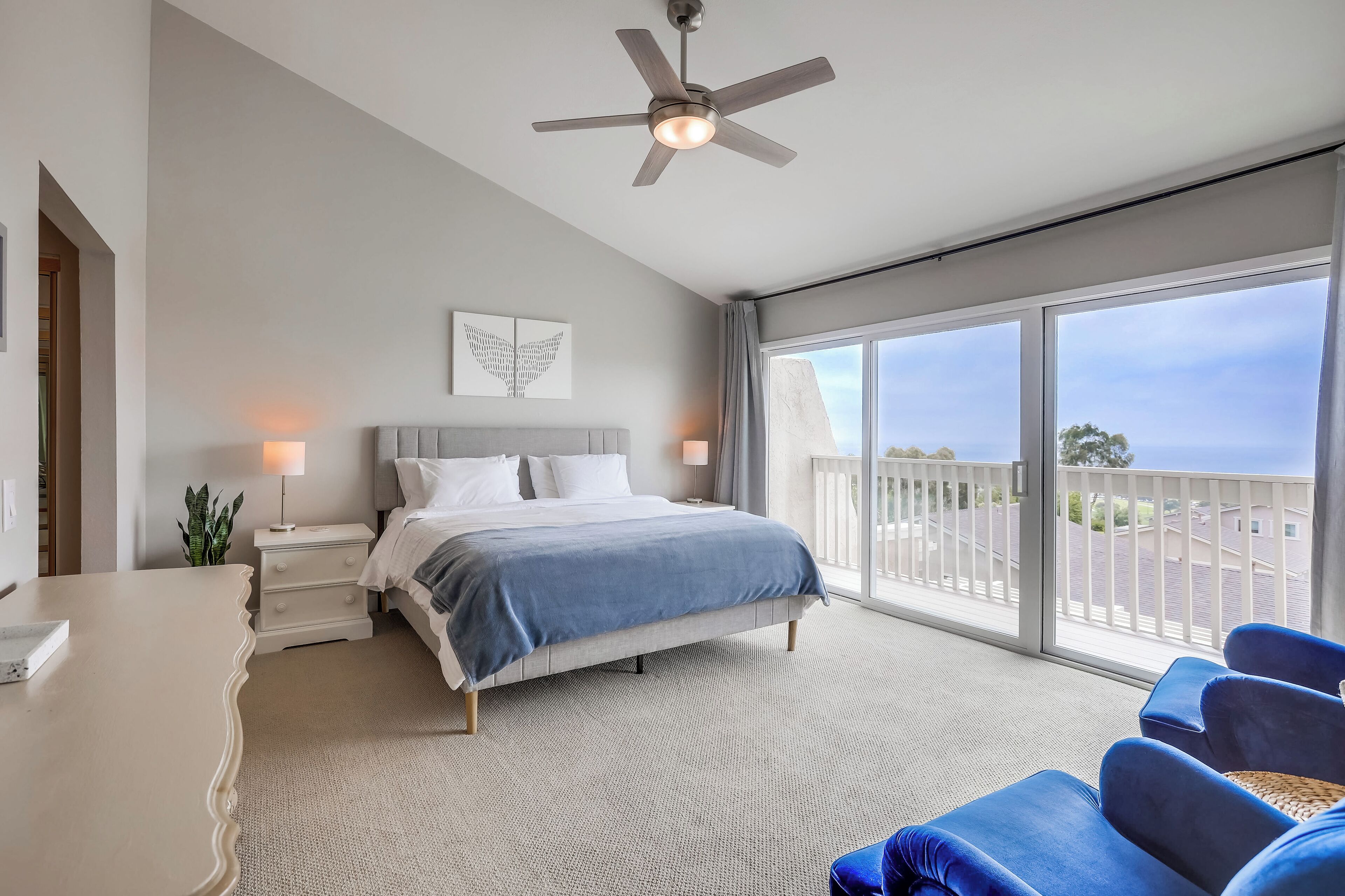 Une chambre à coucher comprenant un lit, des tables de chevet, un ventilateur de plafond, une grande fenêtre avec vue et un balcon.