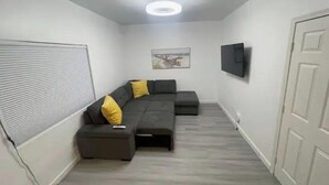 A modern living room with a sectional sofa, a small ottoman, a flat-screen TV mounted on the wall, and a ceiling light fixture. - Beach Rental 3 Blocks to Beach! WIFI, Cable, A/C, and More! (San Diego)