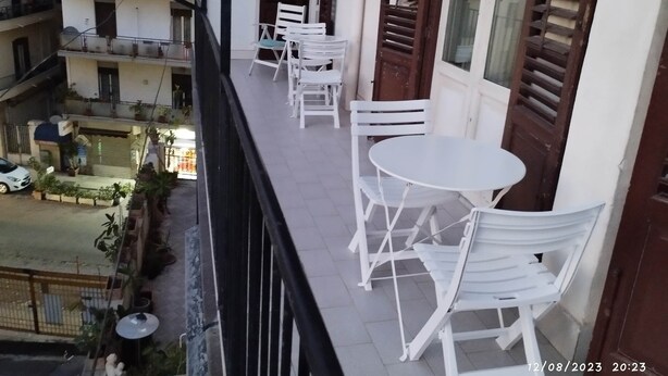 Un balcón con sillas y una mesa blancos, con vista a una calle con edificios y coches aparcados.