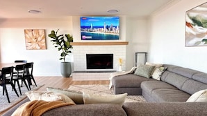Ein modernes Wohnzimmer mit einem Ecksofa, einem Flachbildfernseher über einem Kamin, einem Essbereich mit Stühlen und einer großen Topfpflanze.