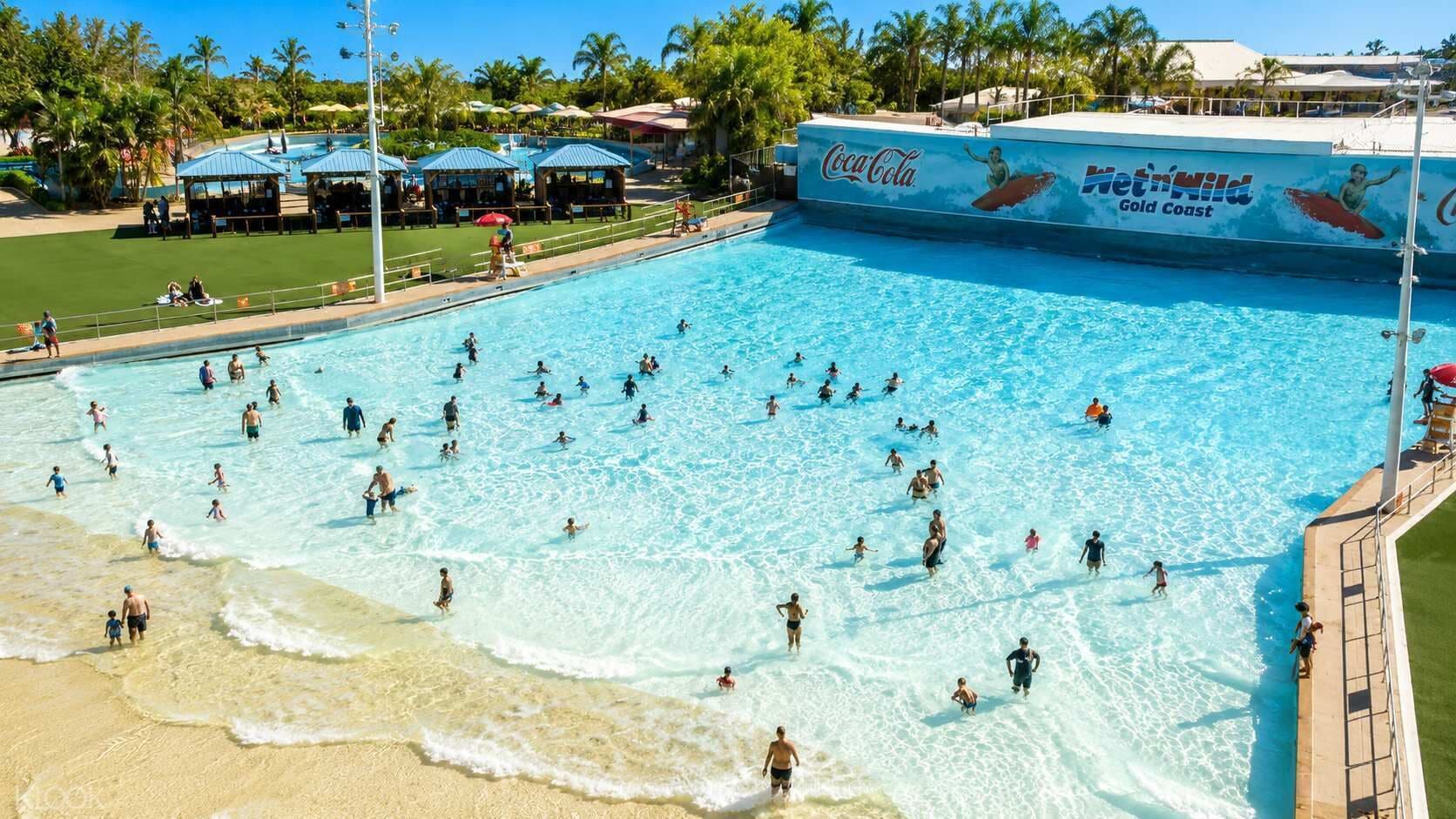 A large swimming pool with people swimming and playing, surrounded by a beach and palm trees.