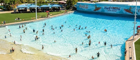 A large swimming pool with people swimming and playing, surrounded by a beach and palm trees.