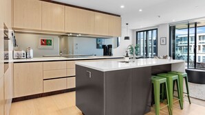 A modern kitchen with wooden cabinets, a large island, green bar stools, and a view of the city through large windows. - Sunny and Spacious in Wynyard Quarter (Auckland)