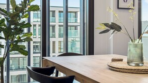 A modern dining area with a wooden table, black chairs, a vase with flowers, and a view of a cityscape through large windows. - Sunny and Spacious in Wynyard Quarter (Auckland)