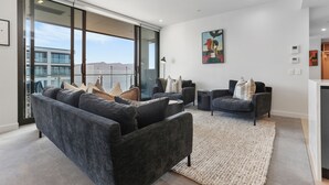A modern living room with a grey sofa, two armchairs, and a coffee table. There is a large window offering a view of buildings and mountains. - Sunny and Spacious in Wynyard Quarter (Auckland)