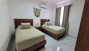 A hotel room with two beds, a nightstand with a lamp, an air conditioning unit, and a window with curtains.