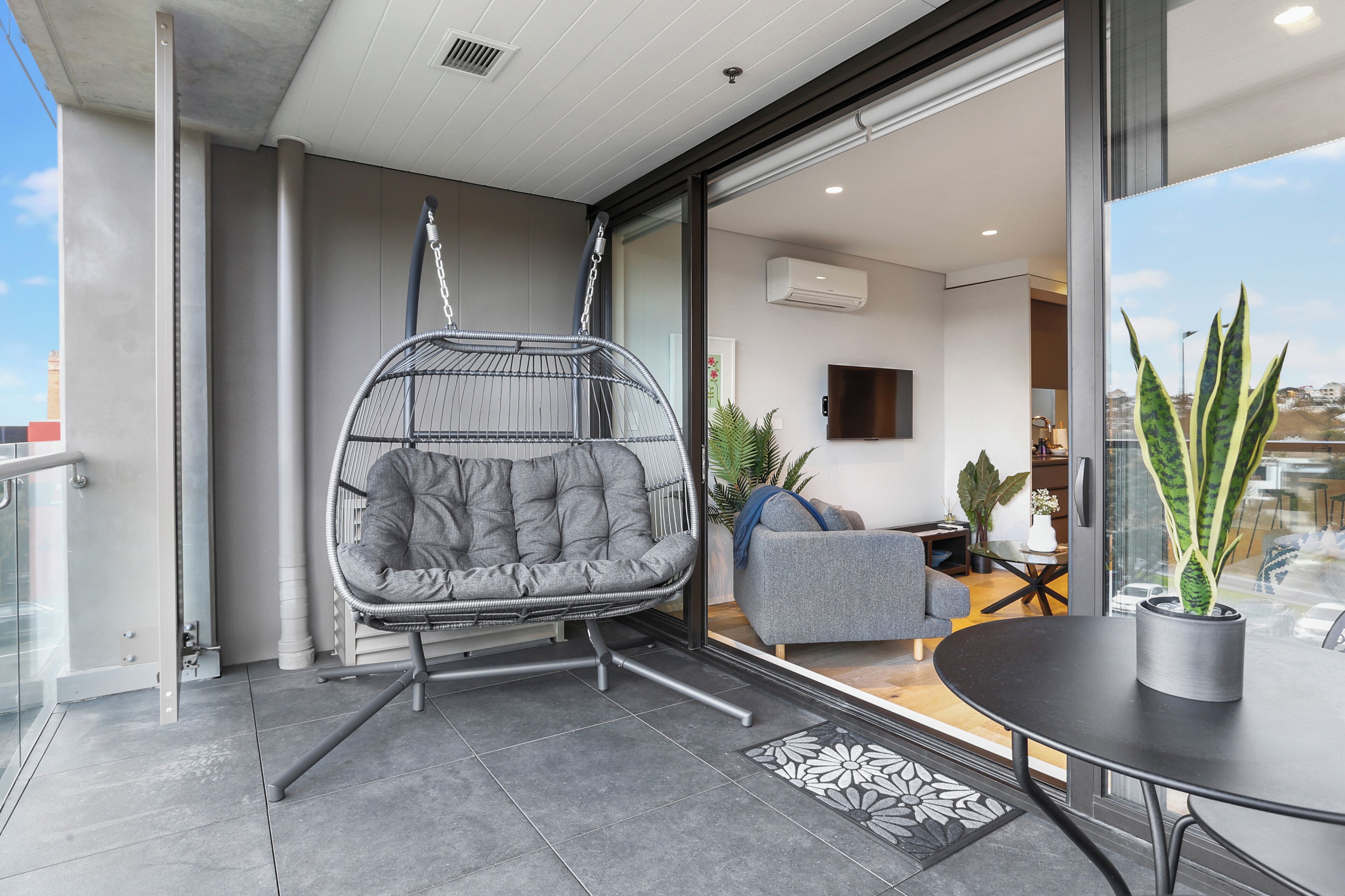 A modern living space with a grey cushioned chair, a small table with a potted plant, and a sofa in the background.