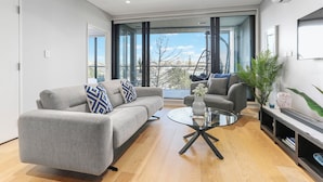 A modern living room with a grey sofa, a glass coffee table, and a view of the outdoors. - Light & relaxing on city fringe (Auckland)