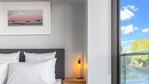 A neatly made bed with white linens, a bedside table with a lamp, and a framed picture of a sunset over water on the wall. - Light & relaxing on city fringe (Auckland)