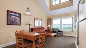 A hotel room with a dining table, chairs, a sofa, a small armchair, and a large window offering a view.