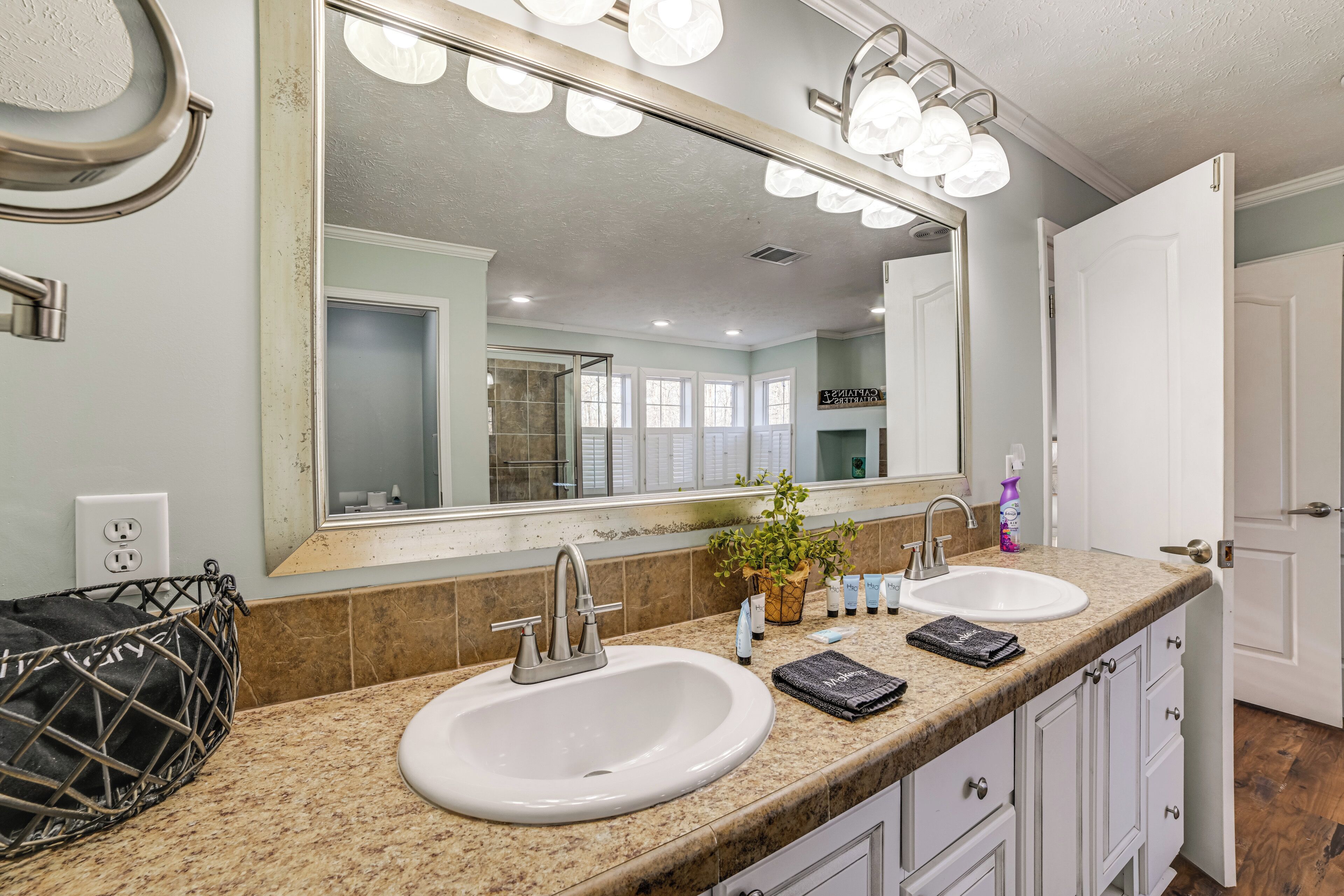 A bathroom with two sinks, a large mirror with lights, a potted plant, and a basket.