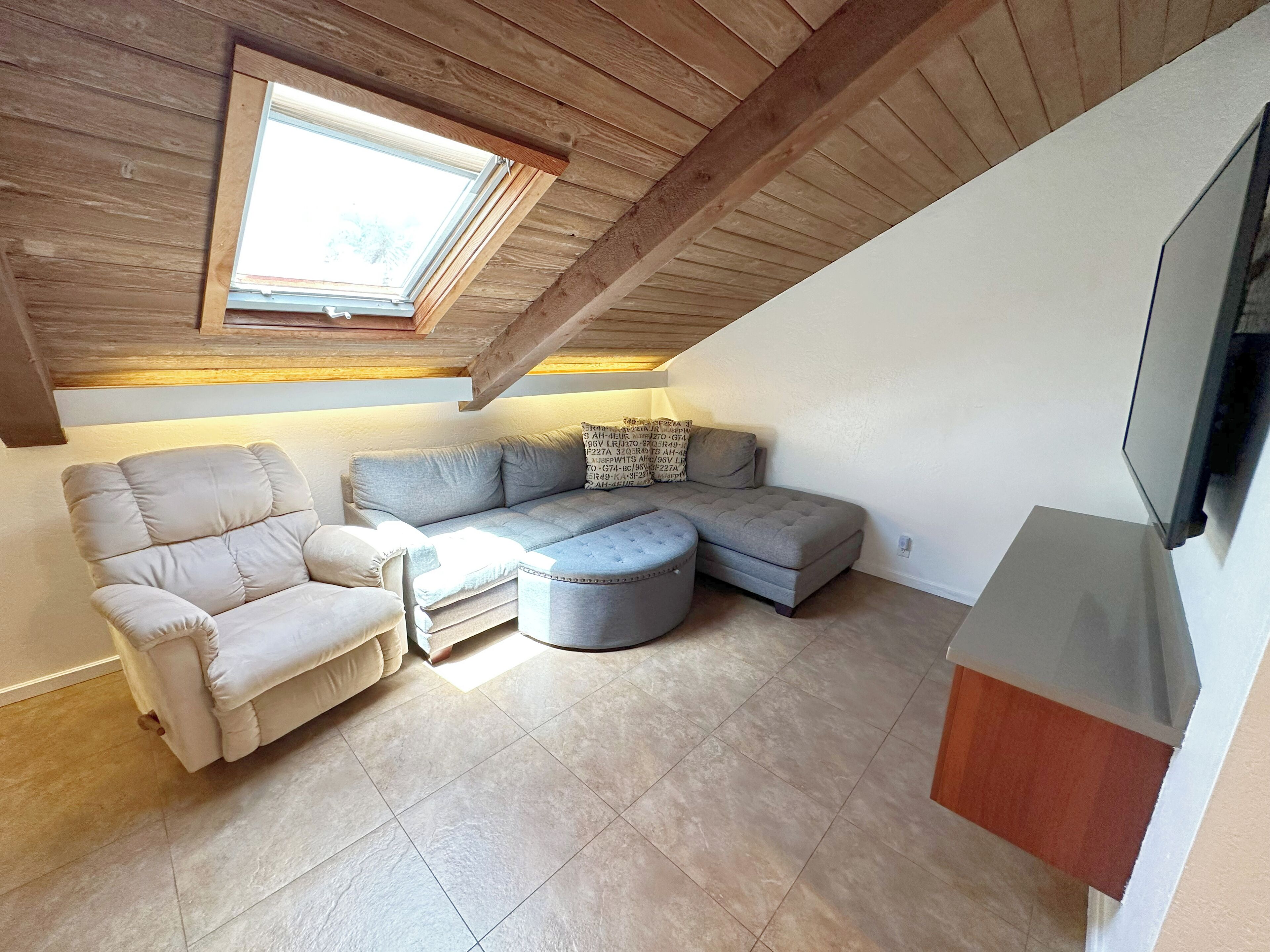 Une pièce mansardée chaleureuse avec un plafond en pente, une grande fenêtre, un canapé d’angle, une chaise longue et un téléviseur à écran plat fixé au mur.