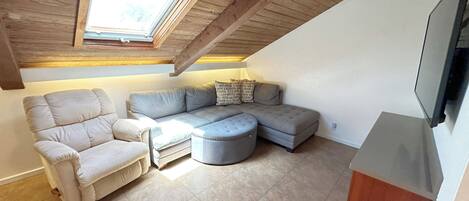 Une pièce mansardée chaleureuse avec un plafond en pente, une grande fenêtre, un canapé d’angle, une chaise longue et un téléviseur à écran plat fixé au mur.