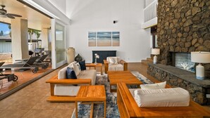 A spacious living area with a stone fireplace, a large window overlooking a beach, and a modern outdoor seating area.