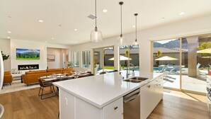 A modern kitchen with a large island, a dining area with a sofa, and a view of a pool and mountains. - Palmer Retreat: #260334 3 BR, Pool, Spa, Foosball! (La Quinta)