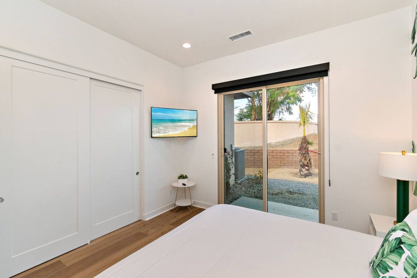 A bedroom with a bed, a small bedside table, a wall-mounted picture, and a sliding glass door leading to an outdoor area.