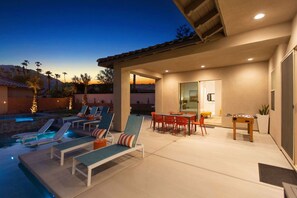 Un área de piscina con sillones de descanso y una mesa, un patio cubierto con una mesa y sillas, y vistas a las montañas al fondo.