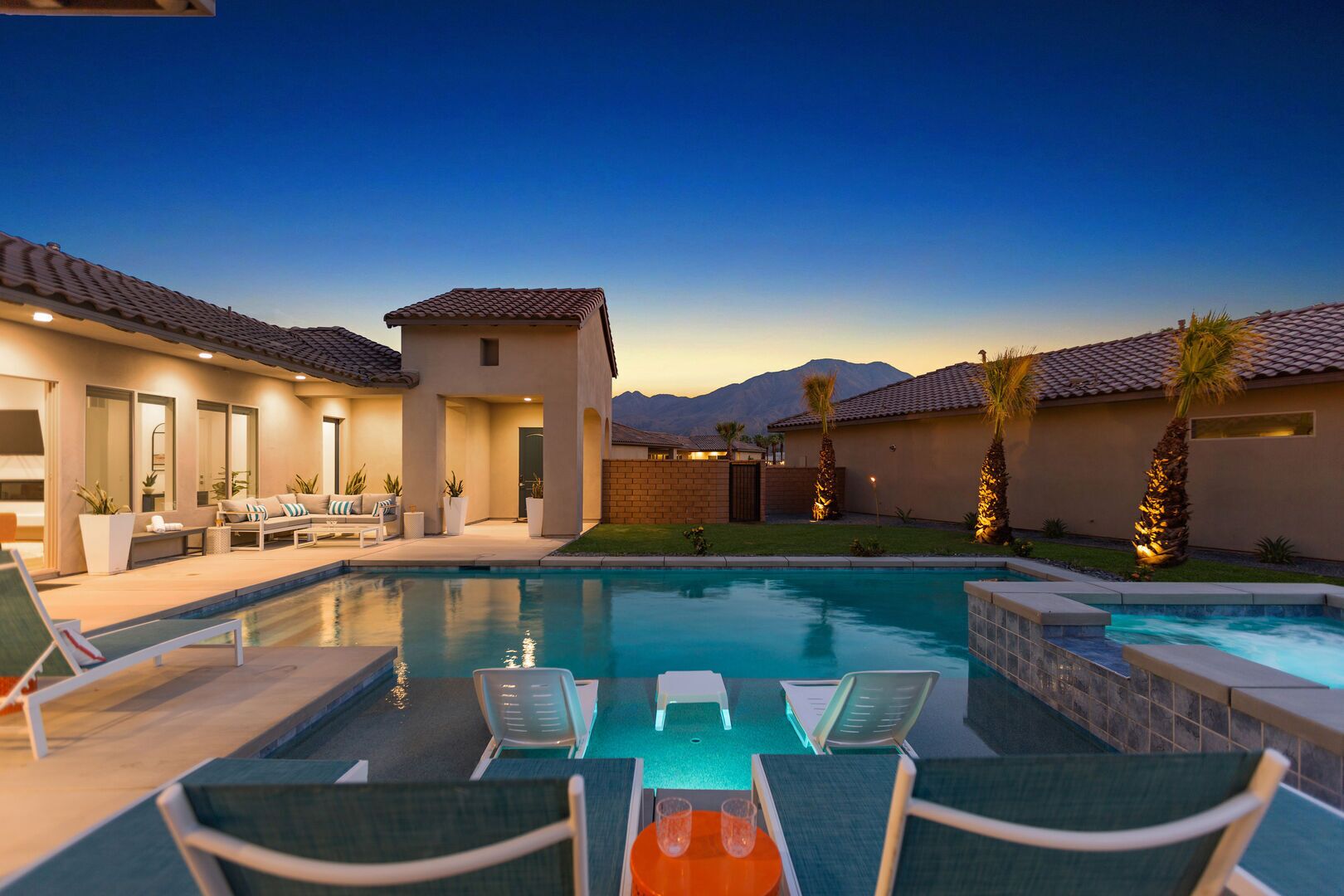 A pool area with lounge chairs and a small building, surrounded by palm trees and mountains in the background.