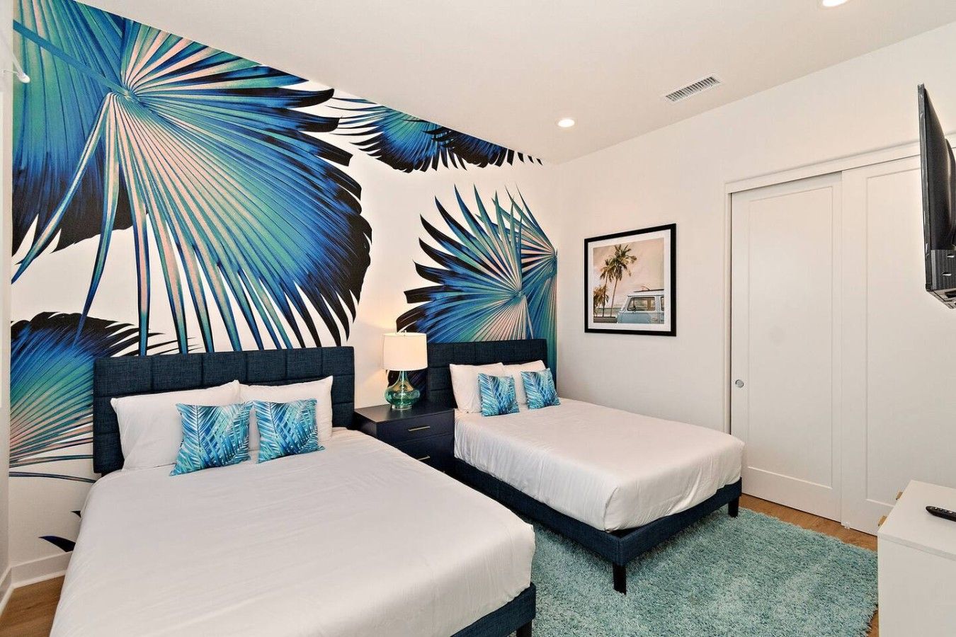 A hotel room with two beds, a large tropical leaf mural, a framed picture on the wall, and a television on the right.