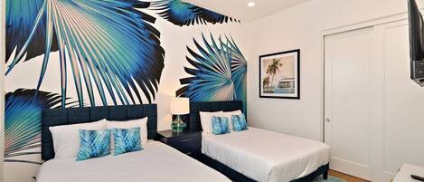 A hotel room with two beds, a large tropical leaf mural, a framed picture on the wall, and a television on the right.