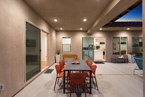 A modern patio area with a dining table and chairs, a sofa, and a small kitchen visible through glass doors. - Palmer Retreat: #260334 3 BR, Pool, Spa, Foosball! (La Quinta)
