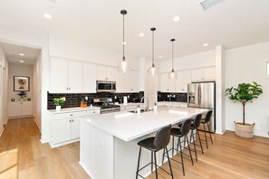 Een moderne keuken met witte kasten, een centraal keukenblok met barkrukken, roestvrijstalen apparatuur en hanglampen.