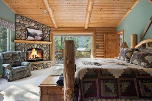 A log cabin bedroom with a stone fireplace, a large bed, a chair, and a view of the outdoors. - Waterview Lodge. New Addition. Spa. Lake views. (Big Bear Lake)