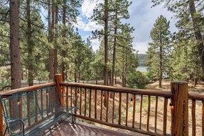 A wooden deck with a metal bench, surrounded by tall pine trees and a view of a lake.