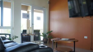 A room with a black leather sofa, a small wooden table, a television mounted on the wall, and a door leading to an outdoor area with a sign. - Private Retreat Getaway (Florence Hall Village)