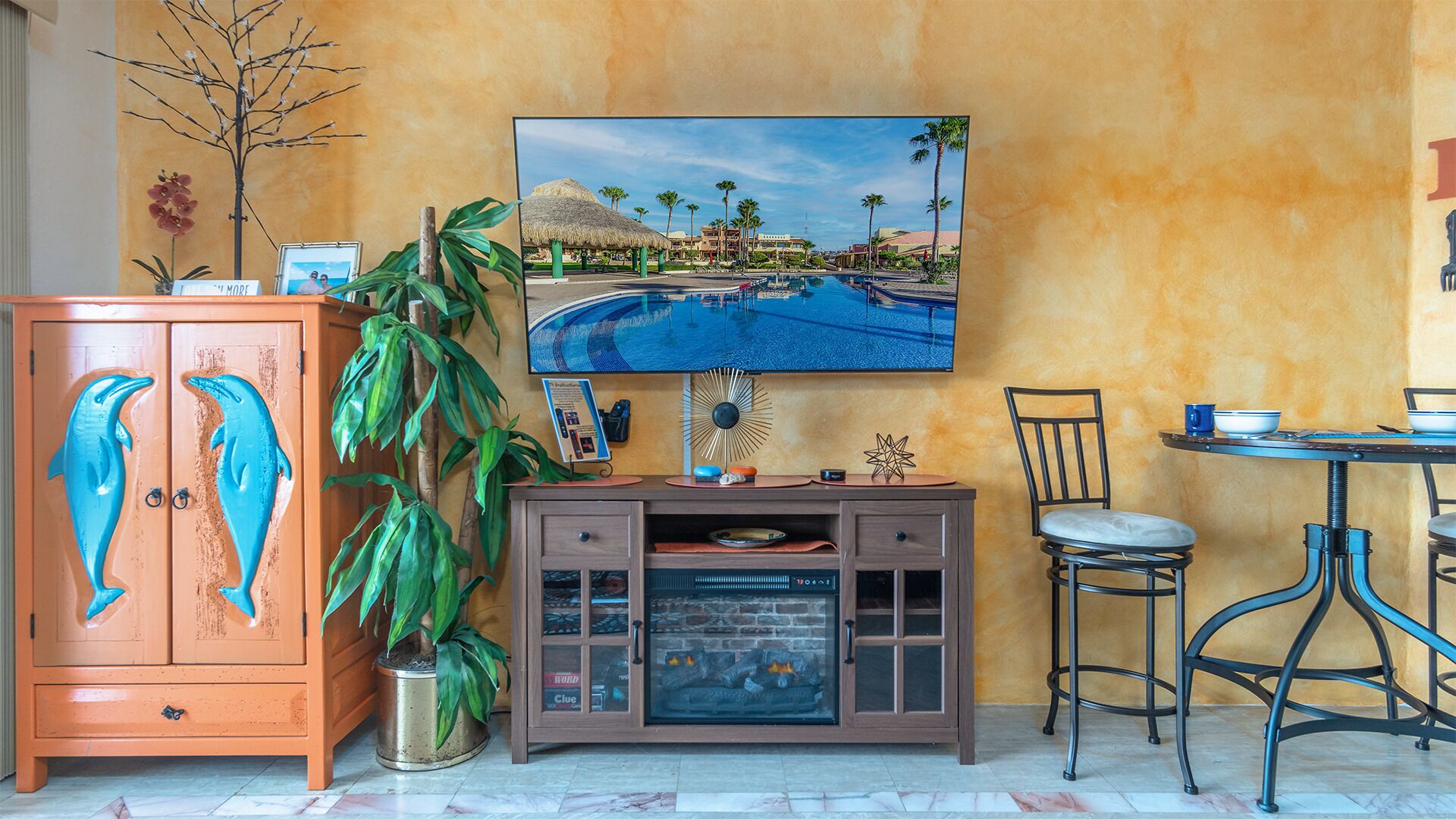 Een woonkamer met een aan de muur bevestigde televisie, een houten kast met dolfijnen decoraties, een grote potplant en een eethoek met een tafel en stoelen.