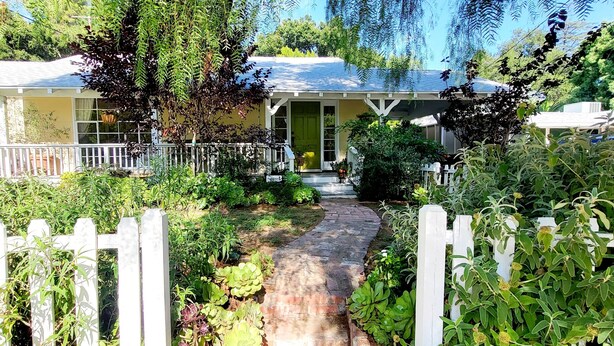 A house with a porch, surrounded by lush greenery and a white picket fence.