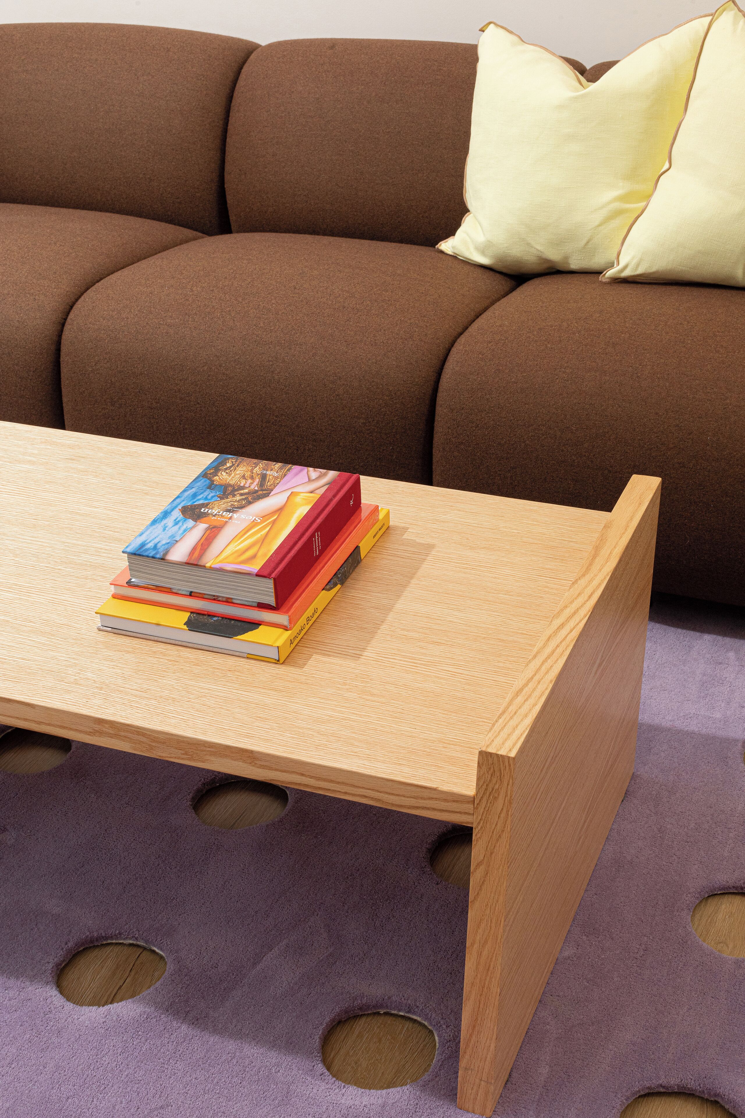 A wooden coffee table with a stack of books on it, placed in front of a brown sofa with a yellow pillow.
