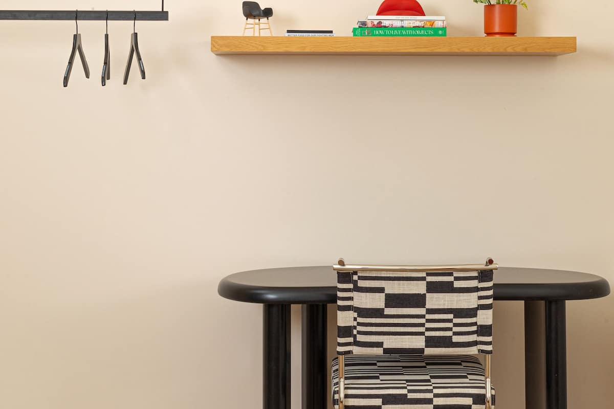 A minimalist room with a wooden shelf holding books, a red cap, and a potted plant. Below, a black table with a striped chair is positioned against a plain wall.