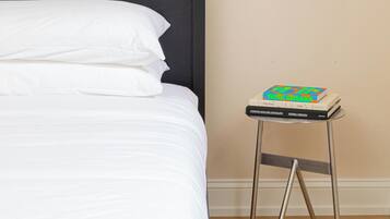 Cama bem arrumada com lençóis brancos, quadro na parede e uma mesinha com livros.