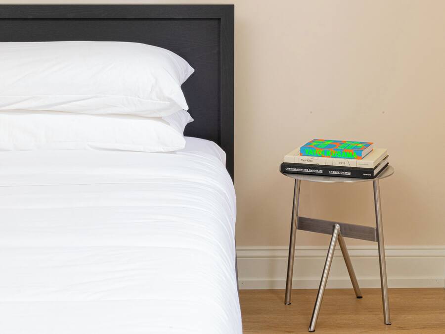 Cama bem arrumada com lençóis brancos, quadro na parede e uma mesinha com livros.