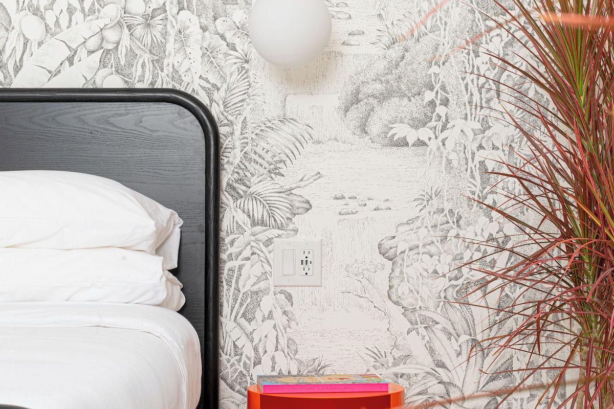 A bedroom with a bed, a wall mural, a hanging light, a small orange storage unit, and a potted plant.