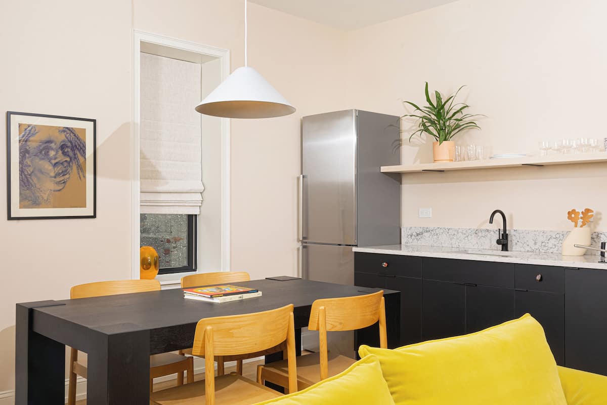 A modern kitchen with a refrigerator, a dining table with chairs, a pendant light, and a potted plant on the counter.
