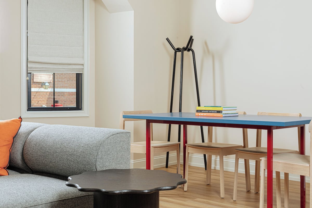 A modern living room with a grey sofa, a small black table, and a blue dining table with red legs. There are two wooden chairs and a floor lamp. A window with blinds is visible, and a stack of books is on the dining table.