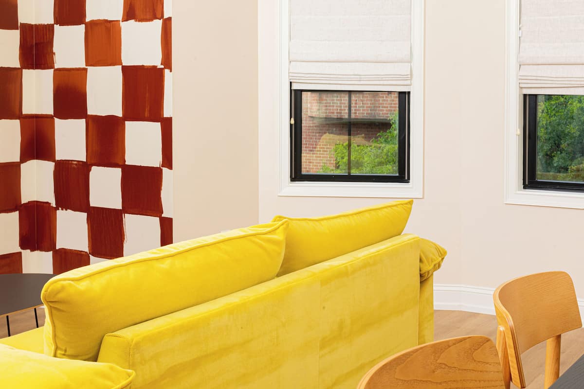 A room with a checkered wall, a yellow sofa, a wooden chair, and windows with blinds.