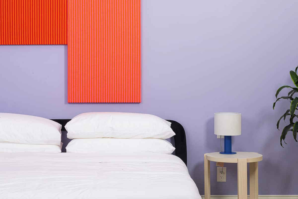 A neatly made bed with white linens, a small bedside table with a lamp, and a potted plant to the right.