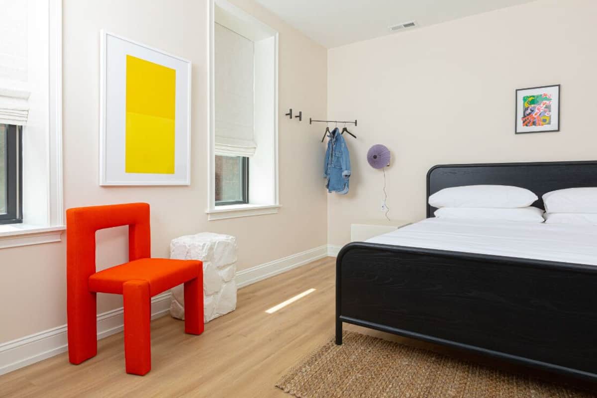 A bedroom with a black bed, a red chair, a white stool, a window with blinds, and a wall with a yellow artwork and a colorful picture.