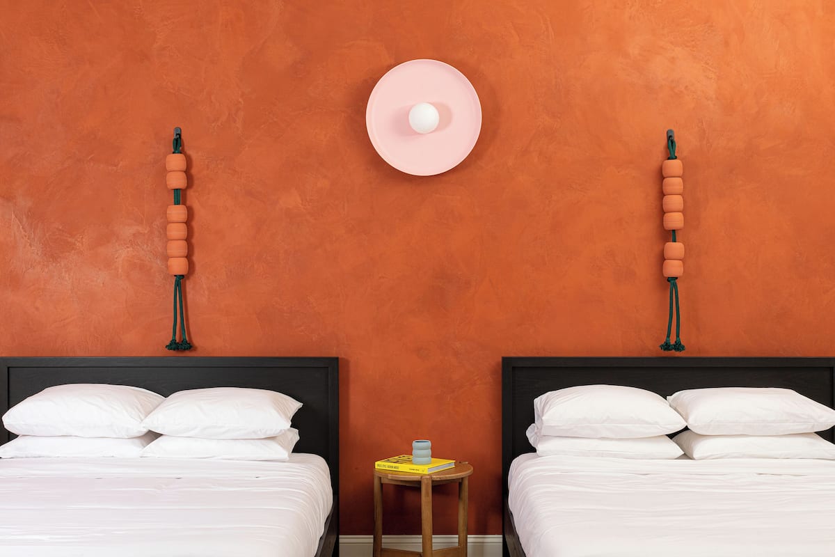 A room with two beds, a small wooden table with a book and a lamp, and a wall-mounted light fixture.