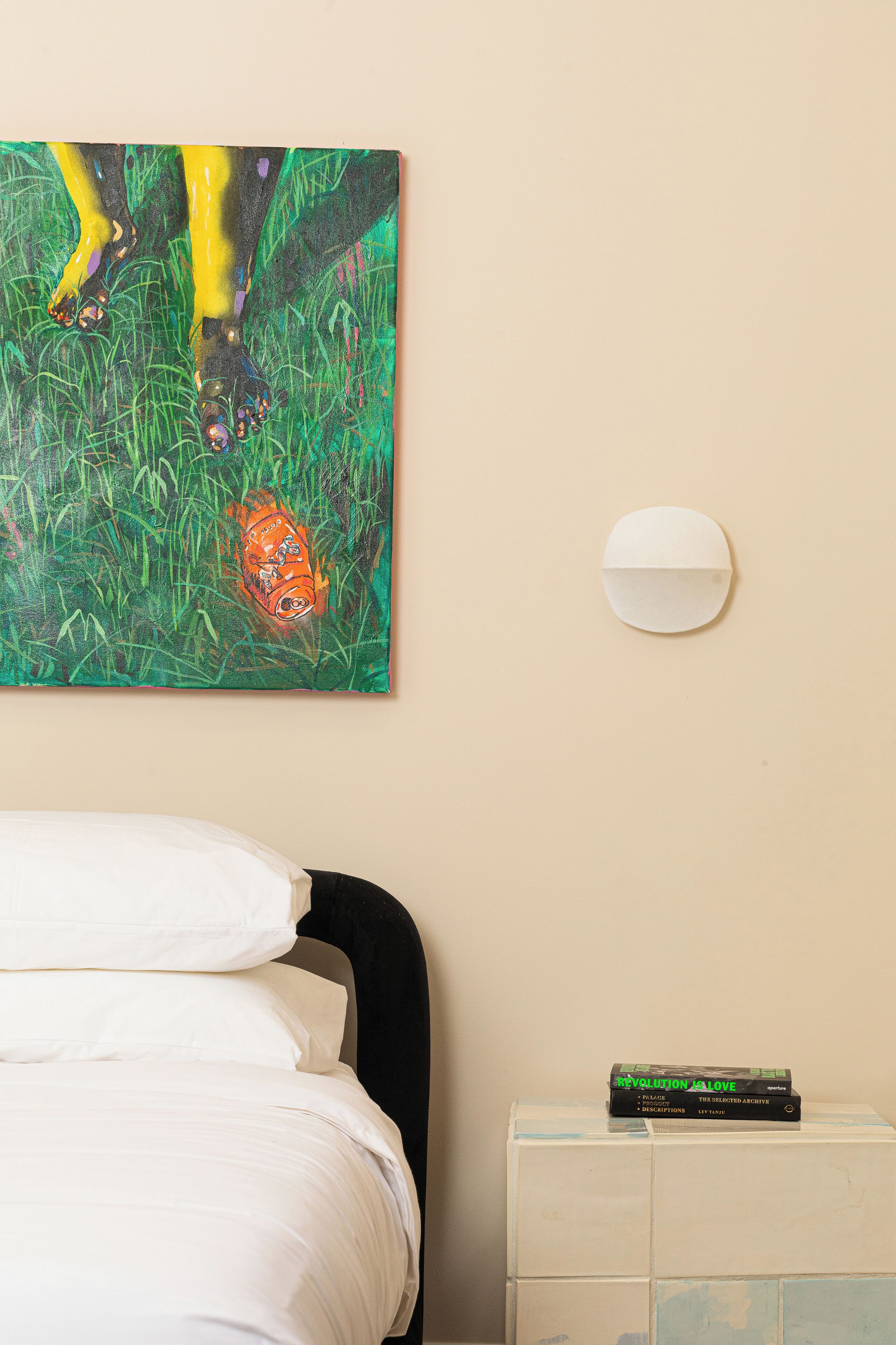 Ein Bett mit weißen Leinen, ein Gemälde einer Gestalt im grünen Gras, eine wandmontierte weiße Lampe und ein kleiner weißer Schrank mit einem Buch darauf.