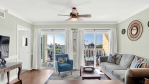 A spacious living room with a large sofa, a blue armchair, and a coffee table. There is a ceiling fan, a TV, and a view of the outside through large sliding doors. - Great Beach Condo, Across From Heated Pool Access (Tybee Island)